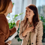 Two young adult businesswomen enjoying a lighthearted conversation in a modern office environment, expressing happiness and connection with genuine warm smiles