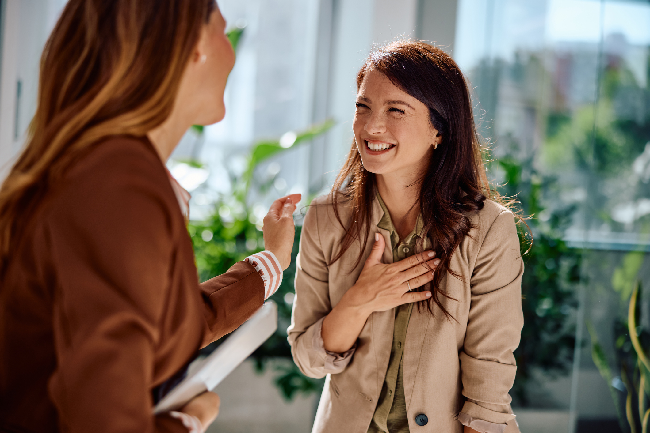 Two young adult businesswomen enjoying a lighthearted conversation in a modern office environment, expressing happiness and connection with genuine warm smiles